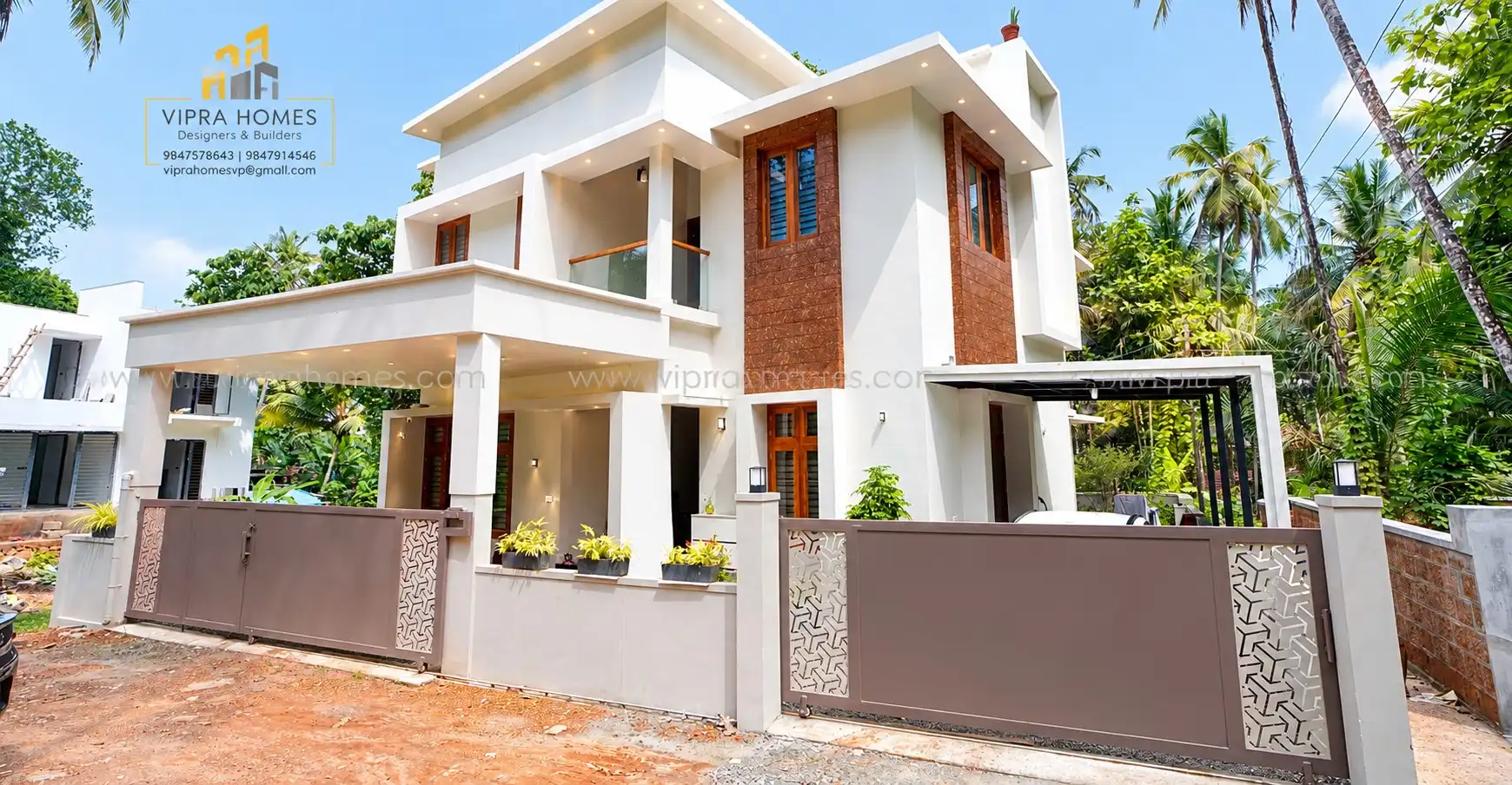 A white two-storey VIPRA HOMES residence with a laterite-brick tower, car porch and patterned gate